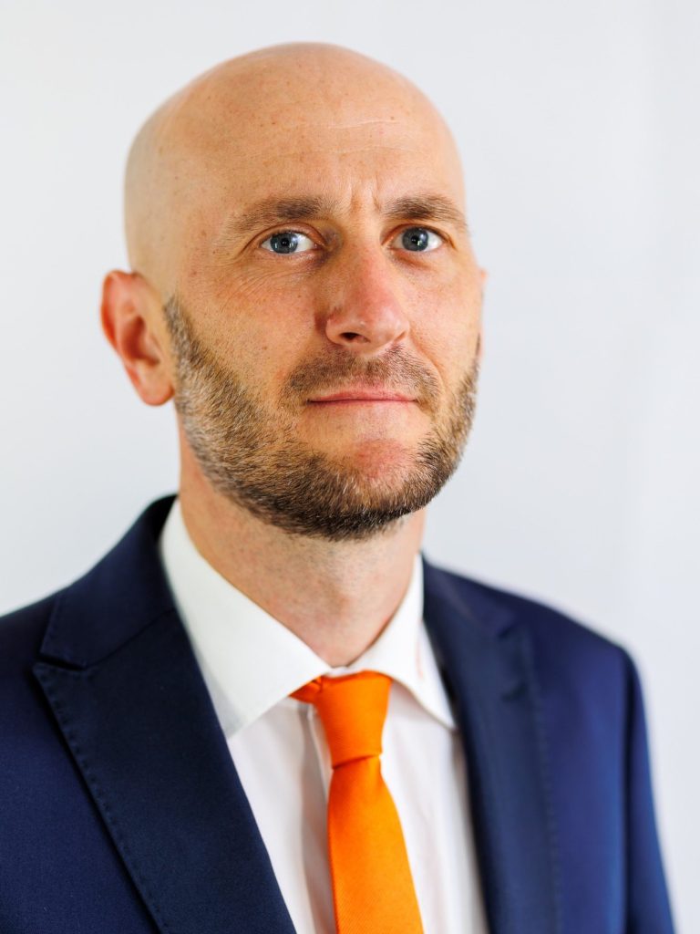 Porträt eines Mannes mit kurzem, glattem Haar und einem leichten Bart, in einem weißen Hemd.